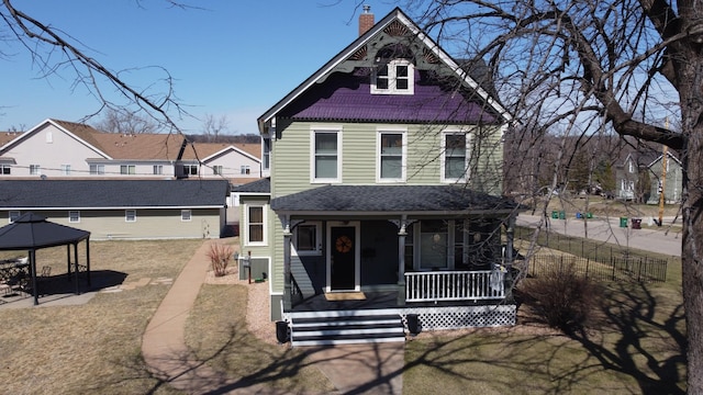 victorian house featuring covered porch, a chimney, roof with shingles, a front lawn, and a gazebo