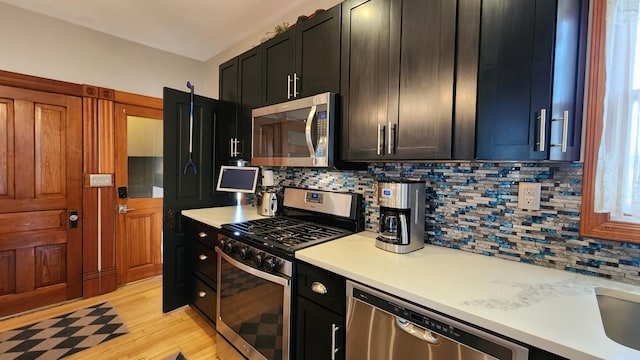 kitchen featuring light countertops, dark cabinetry, decorative backsplash, light wood finished floors, and appliances with stainless steel finishes