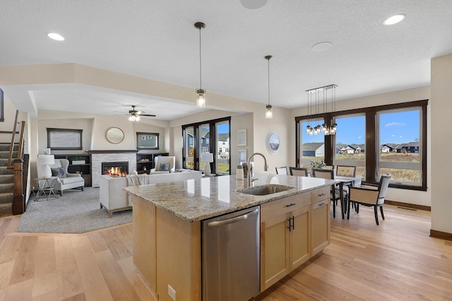 kitchen with a kitchen island with sink, a stone fireplace, light wood-type flooring, stainless steel dishwasher, and light stone counters