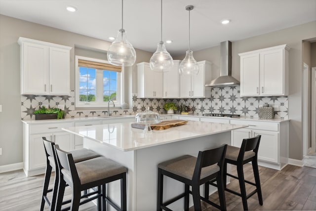 kitchen with wall chimney range hood, an island with sink, wood-type flooring, backsplash, and white cabinetry