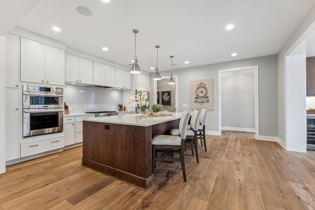 kitchen with white cabinets, a center island with sink, hanging light fixtures, light wood-type flooring, and double oven