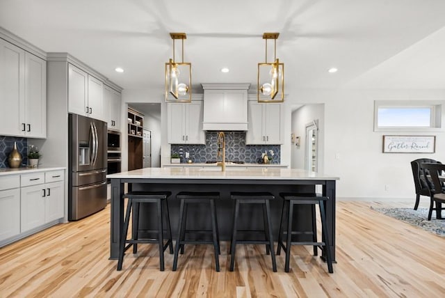 kitchen with stainless steel fridge, custom range hood, a center island with sink, decorative light fixtures, and a breakfast bar area