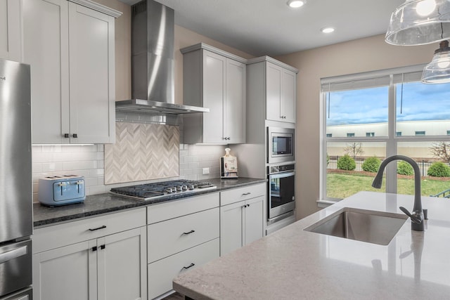 kitchen with wall chimney range hood, a sink, dark stone countertops, appliances with stainless steel finishes, and backsplash