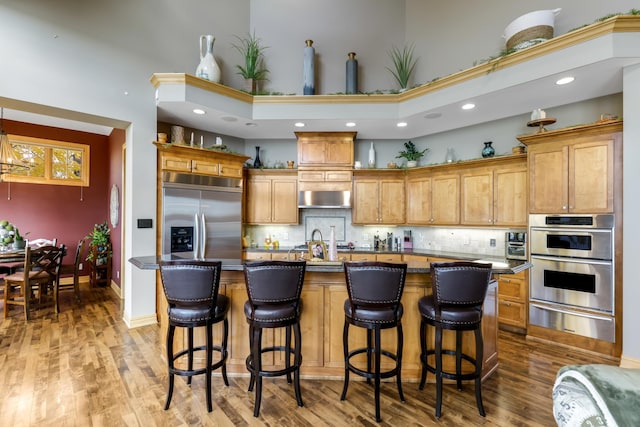 kitchen with hardwood / wood-style flooring, appliances with stainless steel finishes, backsplash, an island with sink, and range hood