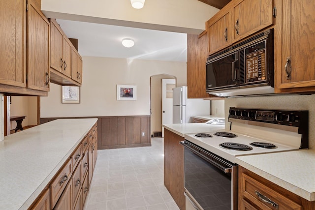 kitchen with a wainscoted wall, white appliances, arched walkways, brown cabinetry, and light countertops
