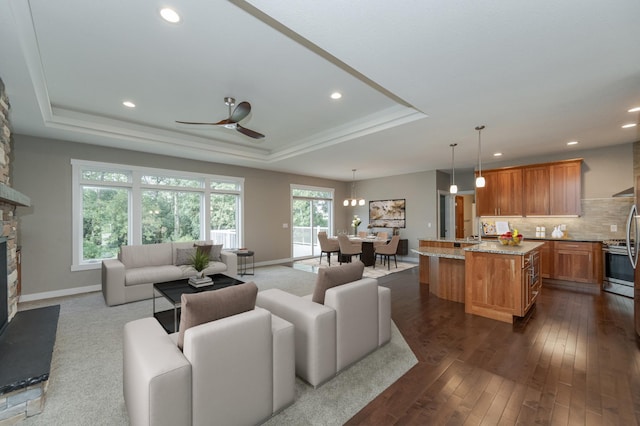 living room with a stone fireplace, ceiling fan with notable chandelier, dark hardwood / wood-style flooring, and a tray ceiling