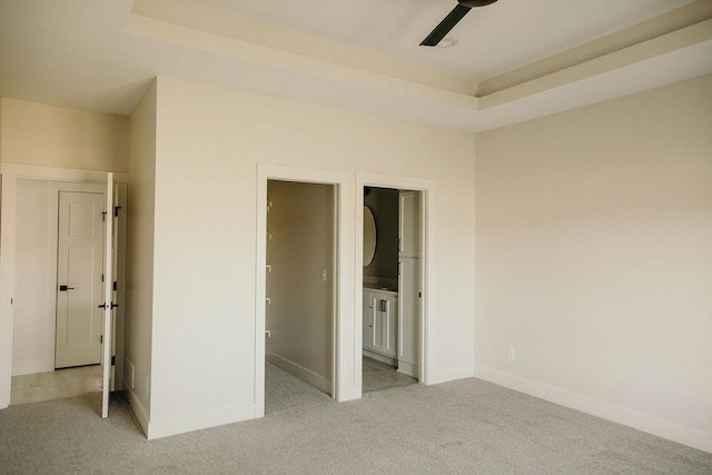 unfurnished bedroom featuring a raised ceiling, ensuite bathroom, ceiling fan, and light colored carpet