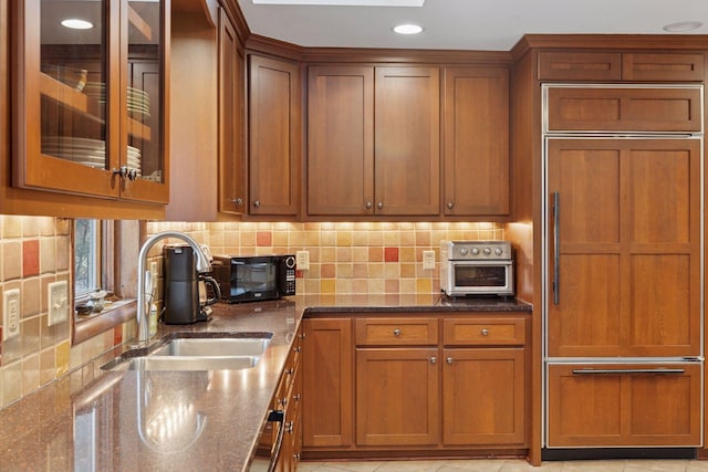 kitchen featuring brown cabinetry, a sink, dark stone countertops, and paneled built in fridge