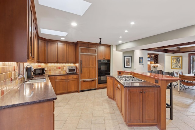 kitchen with dobule oven black, a sink, a kitchen breakfast bar, stainless steel gas stovetop, and a skylight