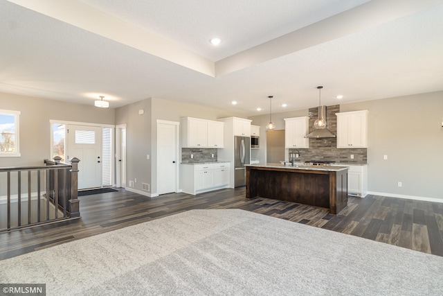 kitchen featuring wall chimney exhaust hood, hanging light fixtures, a center island with sink, stainless steel fridge, and white cabinets