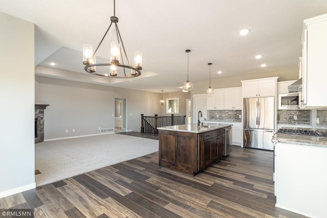 kitchen with a stone fireplace, hanging light fixtures, appliances with stainless steel finishes, a kitchen island with sink, and backsplash