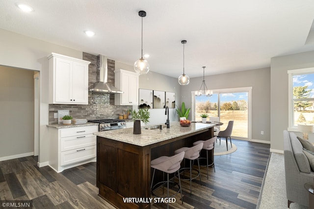 kitchen with wall chimney range hood, decorative backsplash, a sink, dark wood-style floors, and an island with sink