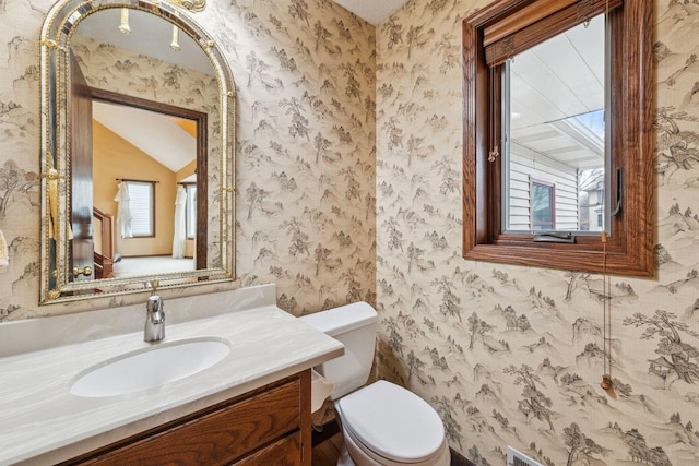 half bathroom with vaulted ceiling, vanity, toilet, and wallpapered walls