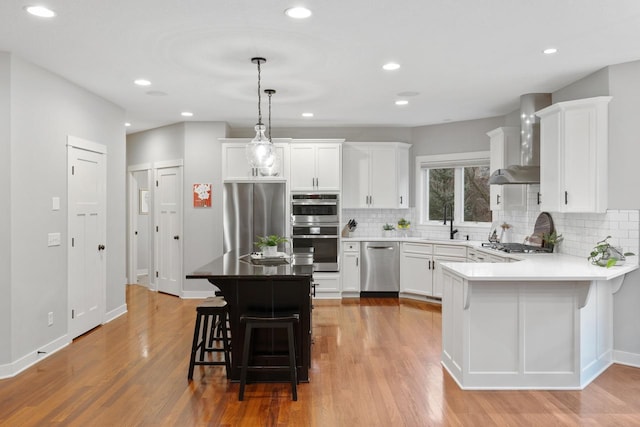 kitchen featuring pendant lighting, wall chimney range hood, white cabinetry, stainless steel appliances, and a breakfast bar