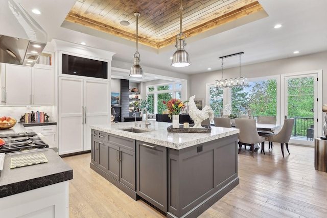 kitchen featuring pendant lighting, a kitchen island with sink, a raised ceiling, sink, and white cabinetry