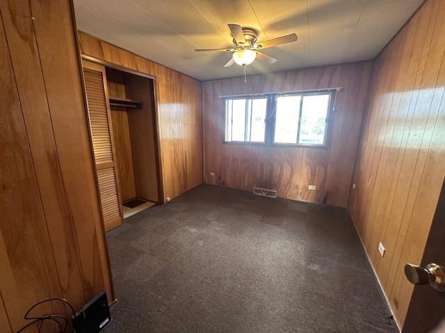 unfurnished bedroom featuring a closet, wooden walls, dark colored carpet, and ceiling fan