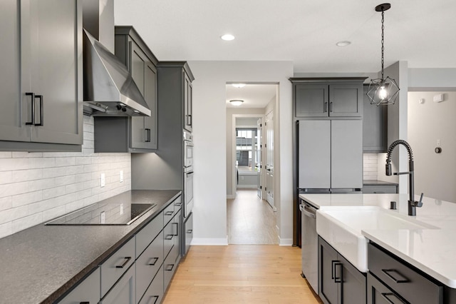 kitchen featuring wall chimney range hood, gray cabinetry, stainless steel appliances, decorative light fixtures, and light wood-type flooring