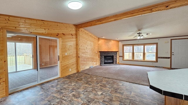 unfurnished living room featuring wood walls, dark carpet, a stone fireplace, ceiling fan, and a textured ceiling