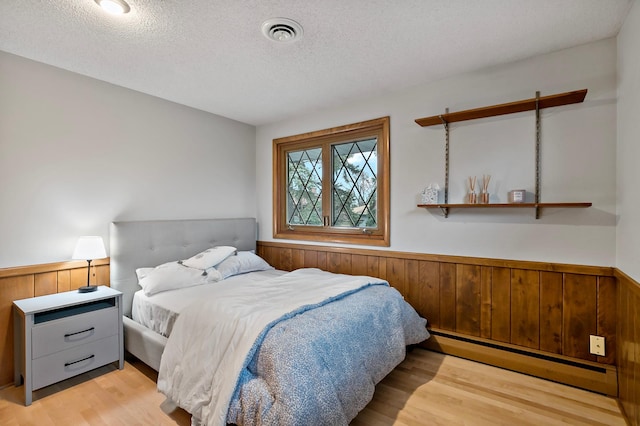 bedroom featuring a textured ceiling, light hardwood / wood-style flooring, wooden walls, and a baseboard radiator