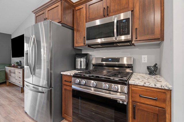 kitchen with light wood-type flooring, stainless steel appliances, brown cabinetry, light stone countertops, and vaulted ceiling