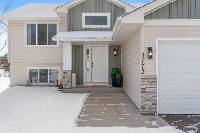 snow covered property entrance with an attached garage, board and batten siding, and stone siding