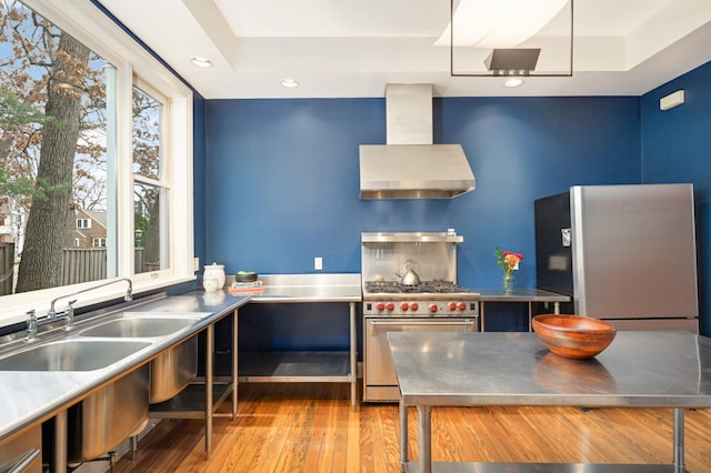 kitchen with stainless steel appliances, sink, stainless steel counters, wall chimney range hood, and a tray ceiling