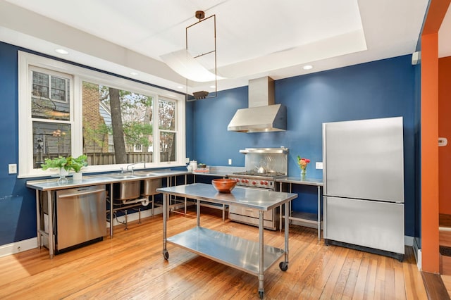 kitchen featuring stainless steel appliances, light wood-type flooring, pendant lighting, wall chimney exhaust hood, and a tray ceiling