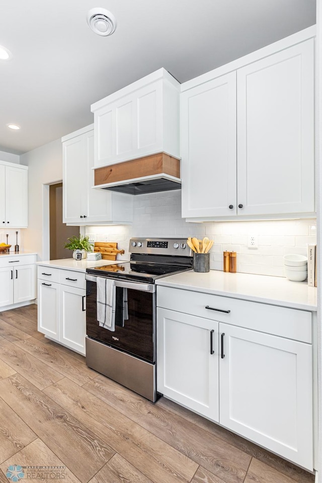 kitchen with stainless steel range with electric stovetop, backsplash, custom range hood, white cabinets, and light wood-type flooring