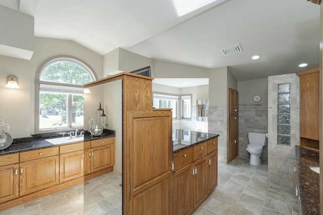 kitchen with tile walls, lofted ceiling, visible vents, brown cabinetry, and dark stone counters