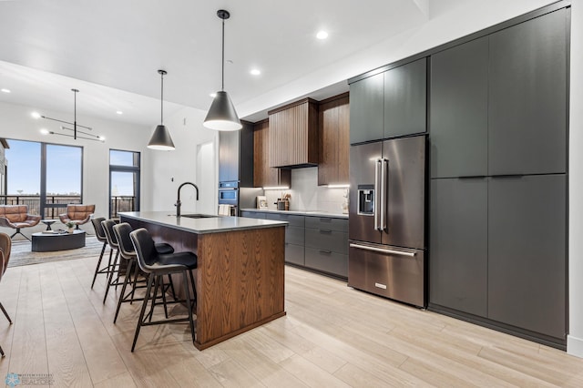 kitchen featuring sink, an island with sink, decorative light fixtures, high end fridge, and light wood-type flooring