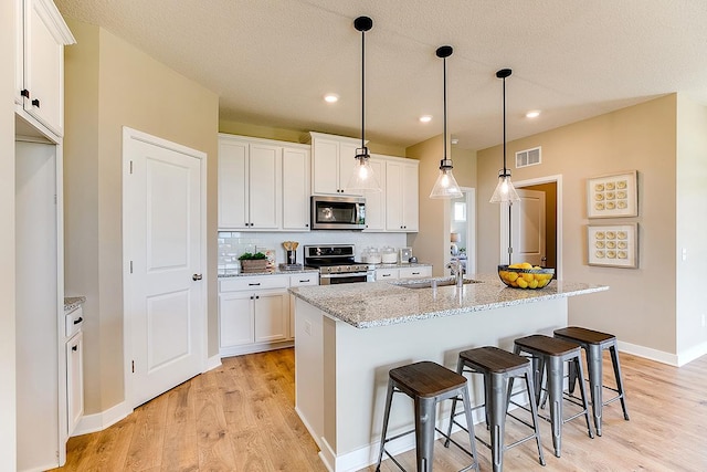 kitchen featuring a kitchen island with sink, white cabinets, appliances with stainless steel finishes, decorative light fixtures, and light stone counters