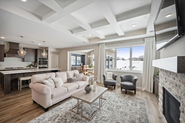 living room with coffered ceiling, a stone fireplace, beamed ceiling, light hardwood / wood-style floors, and a chandelier