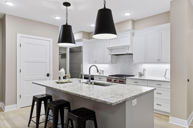 kitchen featuring an island with sink, a sink, built in fridge, light wood finished floors, and decorative backsplash