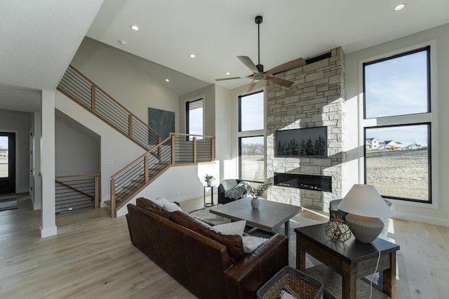 living room featuring ceiling fan, a stone fireplace, a high ceiling, and light wood-type flooring