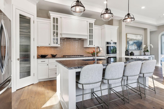 kitchen with white cabinets, a center island with sink, dark stone countertops, appliances with stainless steel finishes, and decorative light fixtures