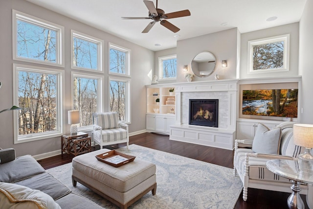 living area with baseboards, dark wood-type flooring, a warm lit fireplace, and ceiling fan