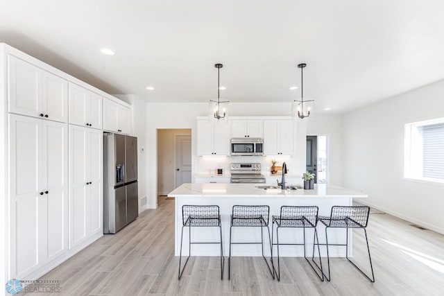 kitchen with stainless steel appliances, light hardwood / wood-style floors, decorative light fixtures, a kitchen island with sink, and white cabinets