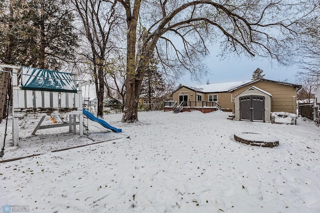 yard layered in snow with a playground, a wooden deck, and a storage unit