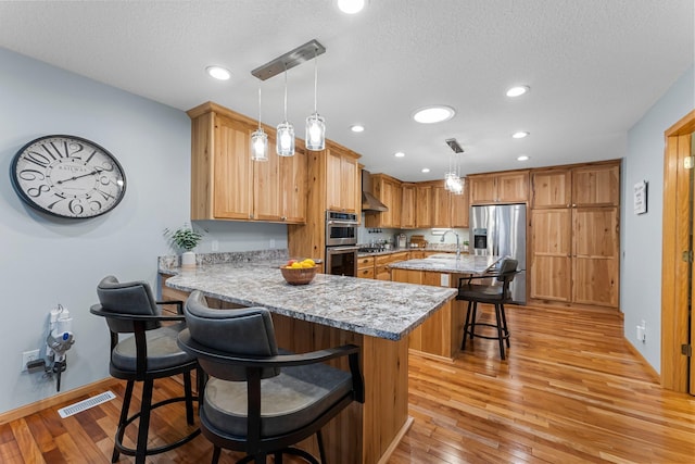 kitchen featuring appliances with stainless steel finishes, decorative light fixtures, a kitchen breakfast bar, kitchen peninsula, and wall chimney exhaust hood