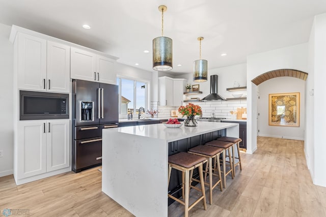 kitchen with wall chimney range hood, high end black refrigerator, white cabinets, a kitchen island, and built in microwave