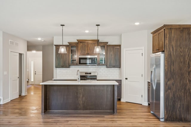 kitchen with glass insert cabinets, light countertops, appliances with stainless steel finishes, a center island with sink, and pendant lighting