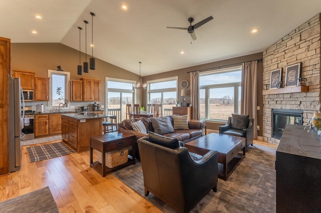 living room featuring ceiling fan with notable chandelier, vaulted ceiling, sink, light hardwood / wood-style flooring, and a stone fireplace