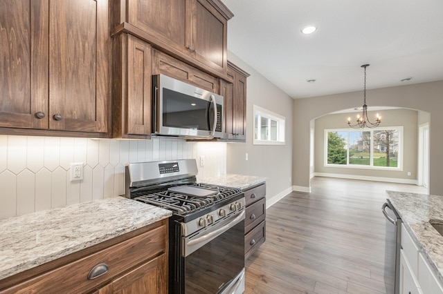 kitchen with light stone countertops, stainless steel appliances, a notable chandelier, backsplash, and light hardwood / wood-style floors