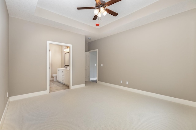 unfurnished bedroom with ceiling fan, ensuite bathroom, light colored carpet, a textured ceiling, and a tray ceiling