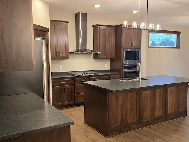 kitchen with sink, appliances with stainless steel finishes, a kitchen island with sink, wall chimney exhaust hood, and light wood-type flooring