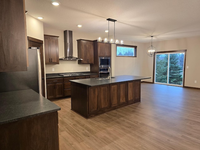 kitchen with wall chimney range hood, appliances with stainless steel finishes, hanging light fixtures, light hardwood / wood-style floors, and a center island with sink