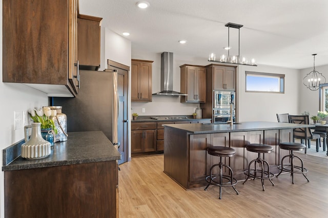 kitchen with wall chimney exhaust hood, a center island with sink, light wood-type flooring, pendant lighting, and stainless steel appliances