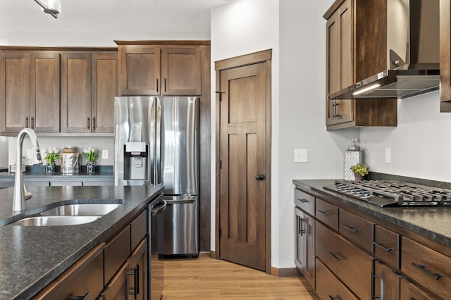 kitchen with wall chimney exhaust hood, sink, dark stone countertops, appliances with stainless steel finishes, and light hardwood / wood-style floors