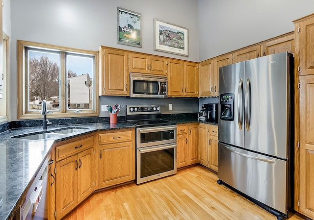 kitchen featuring sink, appliances with stainless steel finishes, dark stone countertops, a towering ceiling, and light wood-type flooring