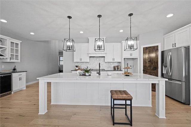 kitchen with wine cooler, stainless steel refrigerator with ice dispenser, light hardwood / wood-style floors, decorative light fixtures, and a kitchen island with sink